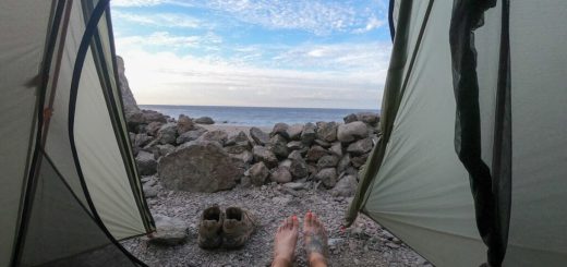 View from inside a tent with a person’s legs and feet visible, overlooking a rocky beach and the ocean under a blue sky with scattered clouds.